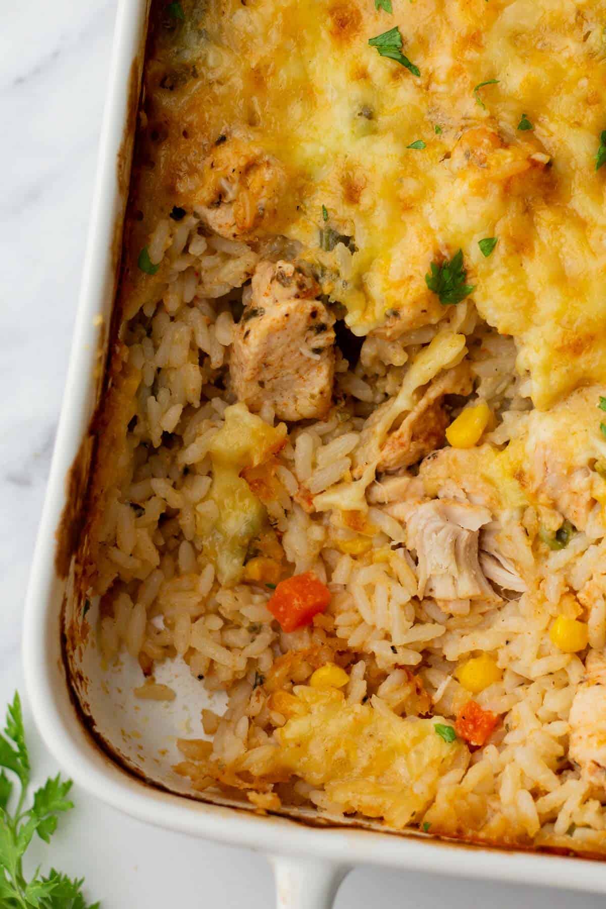 A casserole dish with baked rice, shredded chicken, corn, and melted cheese on top, with a portion served out. Garnished with herbs.