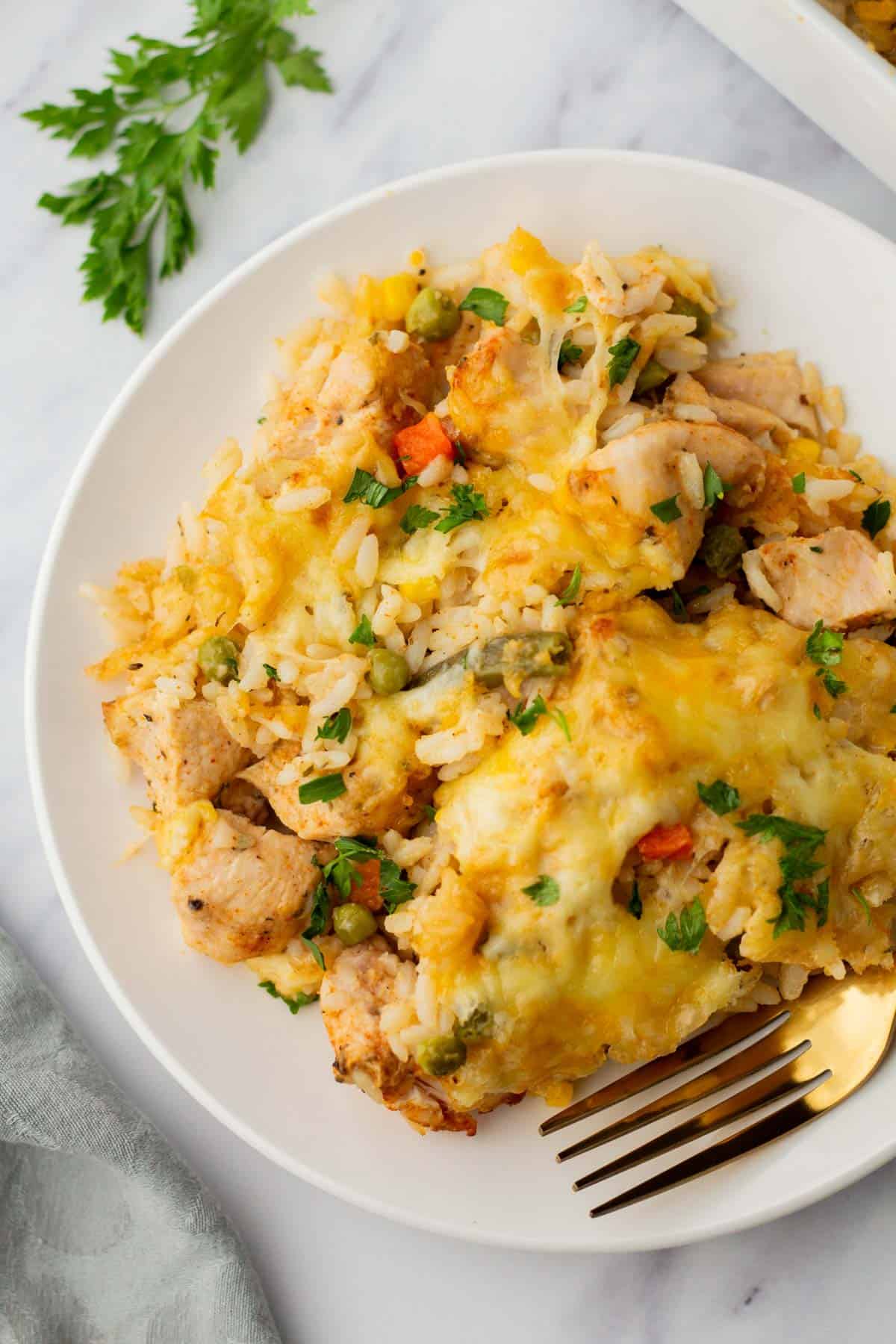 A plate with cheesy chicken and rice casserole topped with herbs. A fork is placed beside the dish on a white surface.