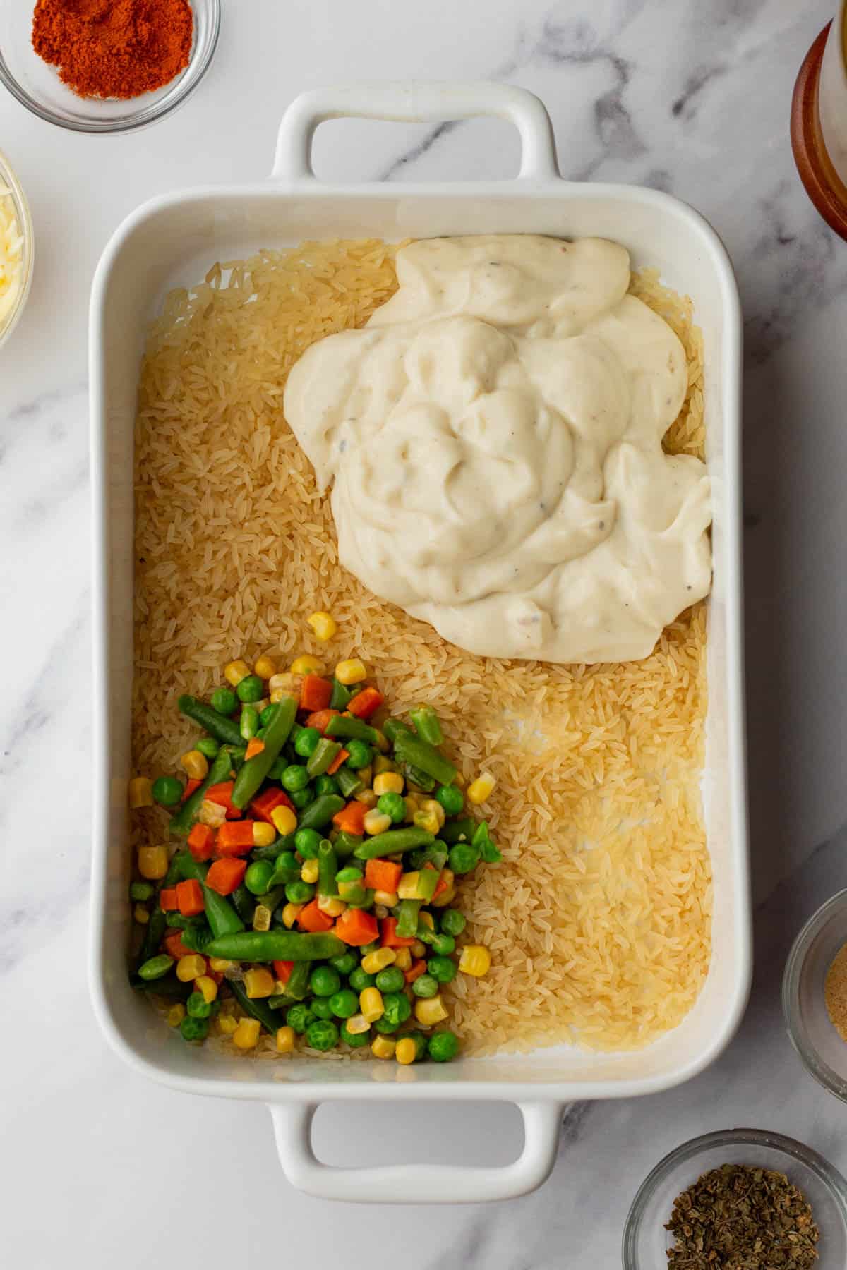 White baking dish with uncooked rice, mixed vegetables, and a dollop of creamy sauce on a marble surface, surrounded by spices in small bowls.