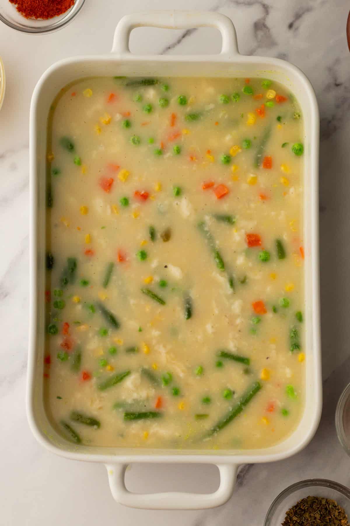 A rectangular dish containing a creamy chicken casserole filling with visible peas, carrots, corn, and green beans.