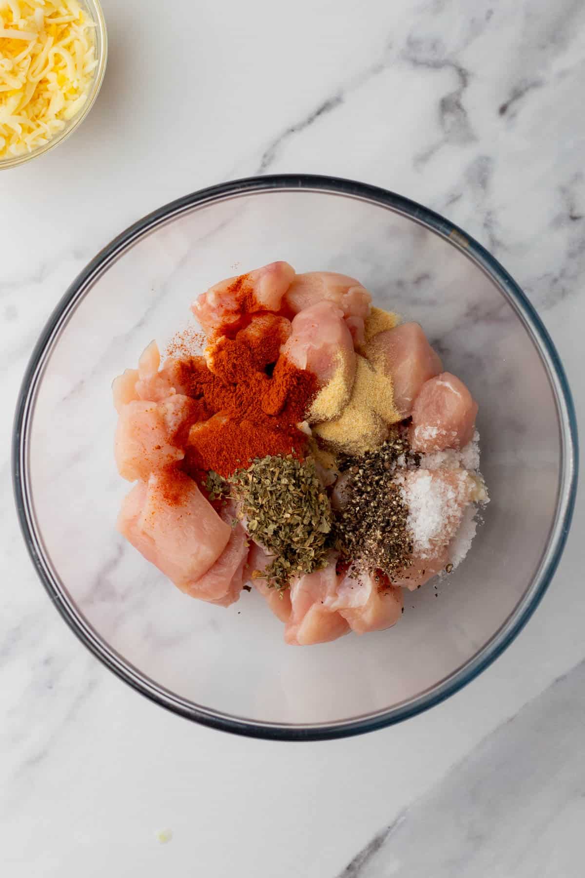 Raw chicken pieces in a bowl with spices and herbs, including paprika, black pepper, garlic powder, oregano, and salt. Shredded cheese is visible in a separate bowl nearby.