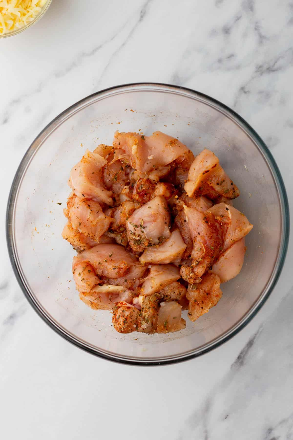 Glass bowl containing seasoned raw chicken pieces on a marble countertop.