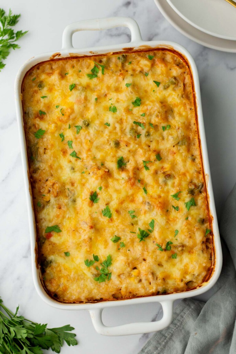 A baked chicken and rice casserole with a golden cheese topping, garnished with chopped parsley, in a white rectangular dish on a marble surface.