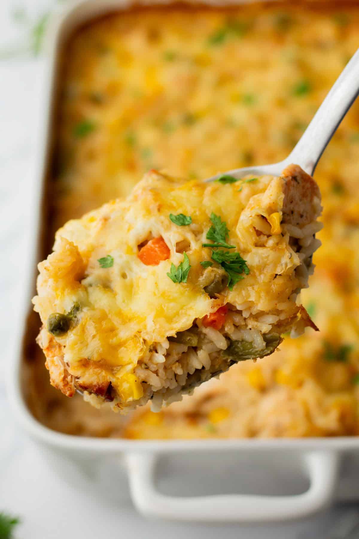 A close-up of a serving spoon lifting a portion of creamy baked chicken and rice casserole with vegetables, topped with melted cheese and garnished with parsley, from a baking dish.