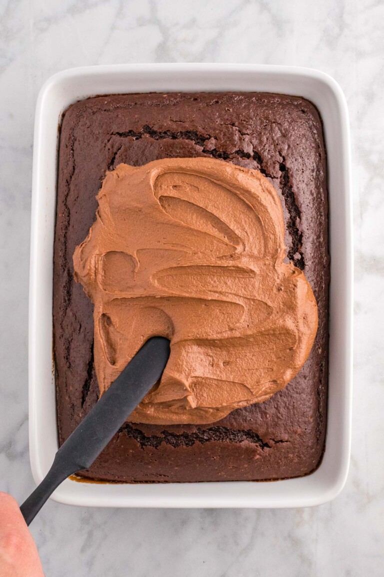 A chocolate sheet cake in a rectangular dish with chocolate buttercream frosting being spread using a spatula.