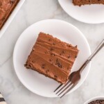 A chocolate cake slice with buttercream frosting and sprinkles on a white plate, accompanied by a fork.