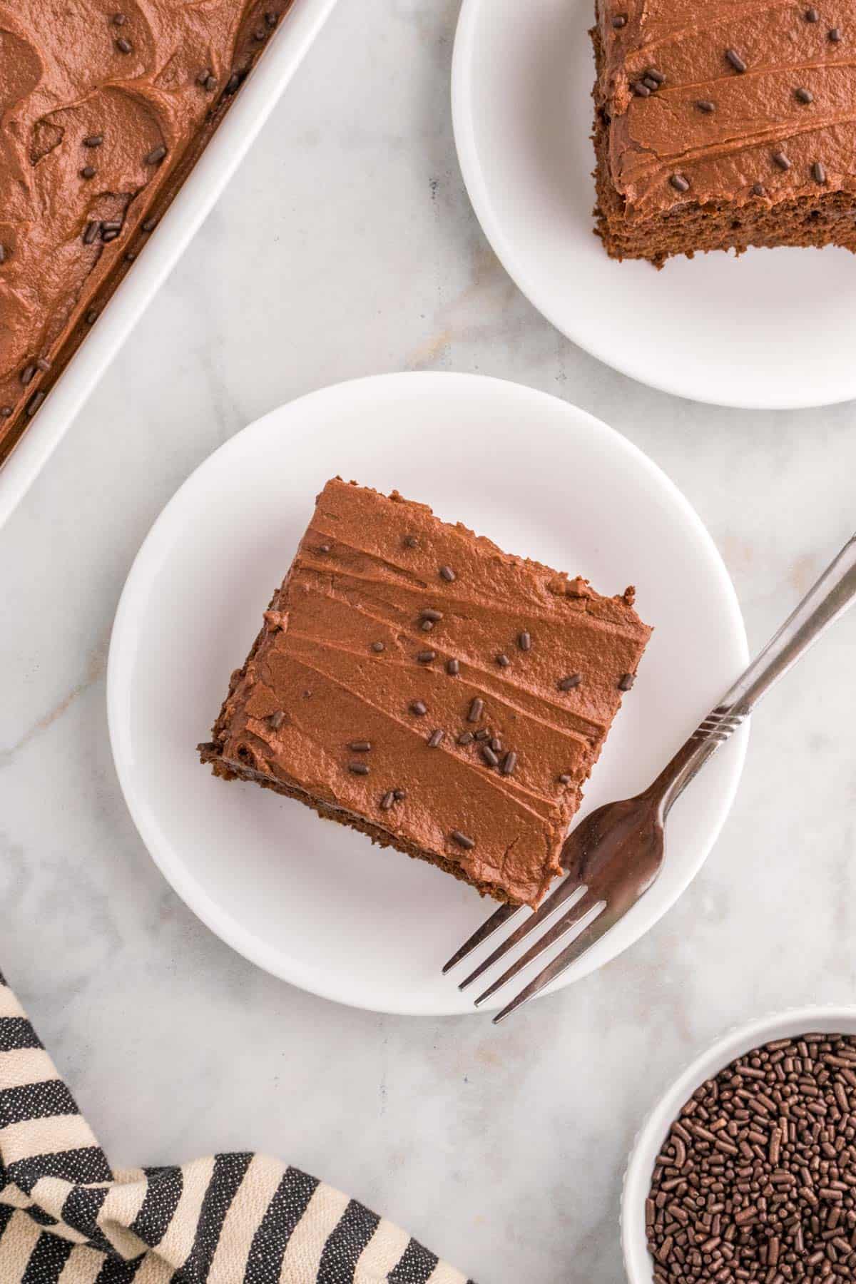 A chocolate cake slice with buttercream frosting and sprinkles on a white plate, accompanied by a fork.