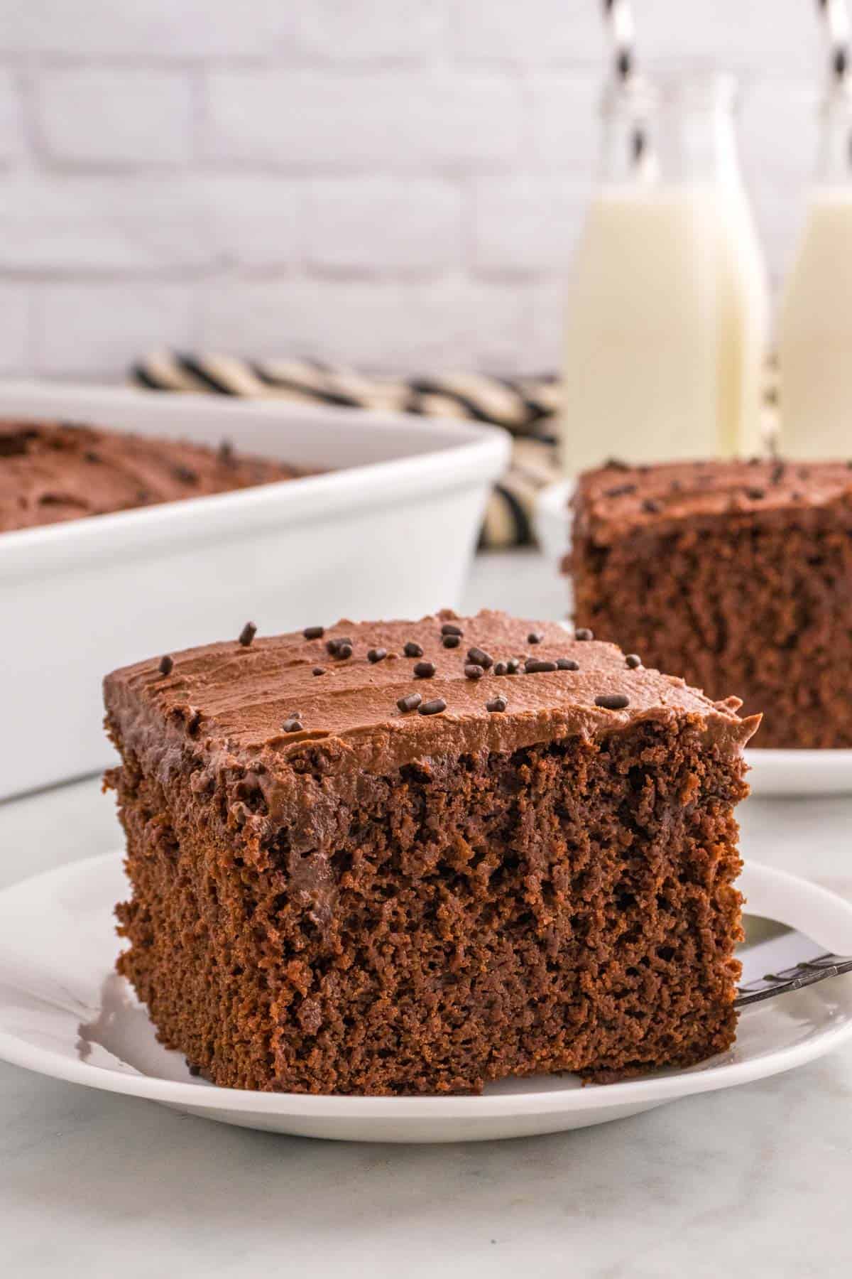 A slice of chocolate cake with buttercream frosting and sprinkles on a plate, with a baking dish and milk bottles in the background.