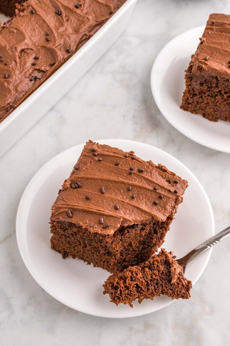 A slice of chocolate cake with chocolate buttercream on a white plate, garnished with chocolate sprinkles, next to a baking dish of more cake. A fork holds a piece of the cake.
