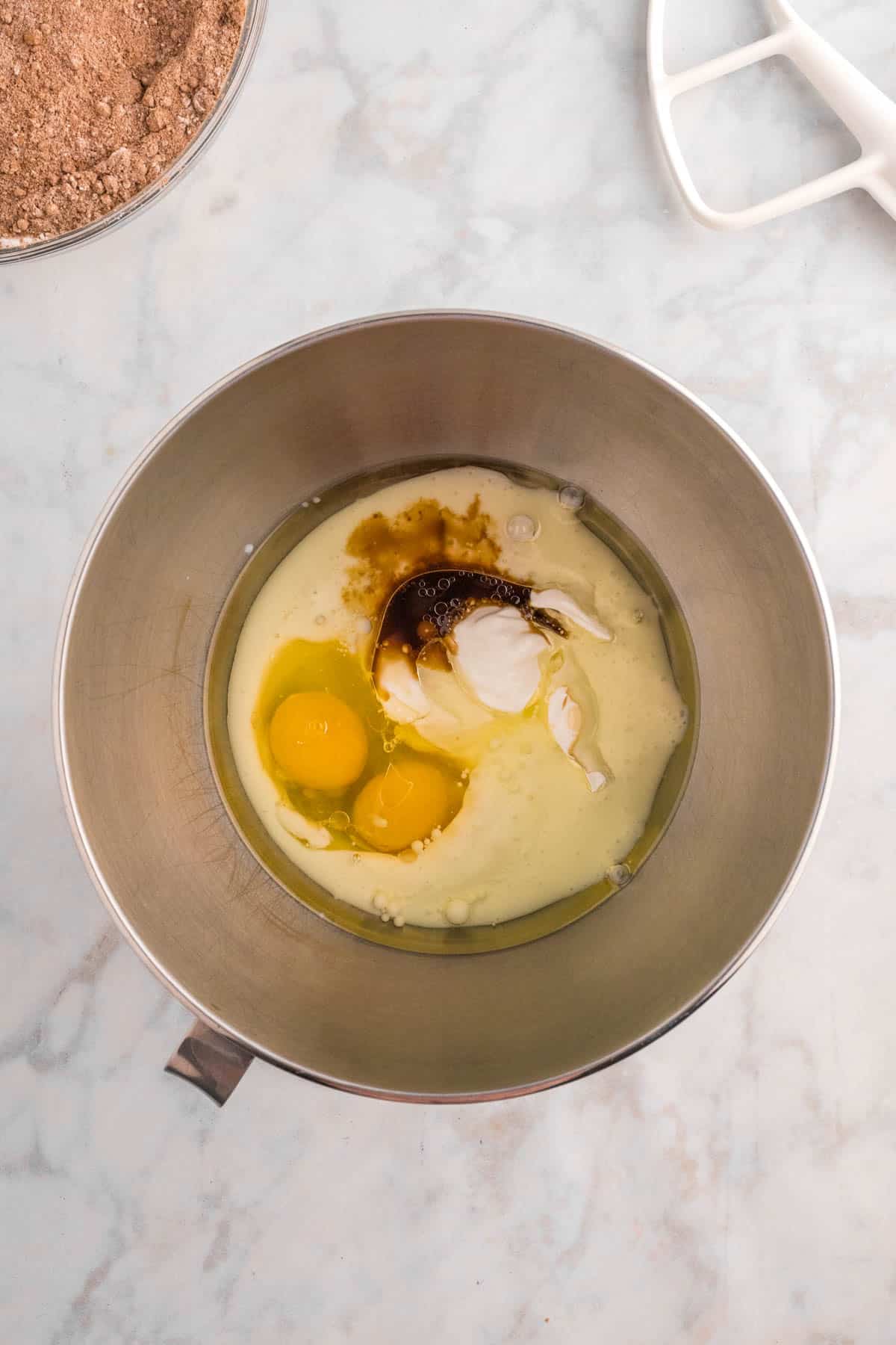 A mixing bowl containing eggs, vanilla, and cream on a marble countertop, with a mixer attachment nearby.