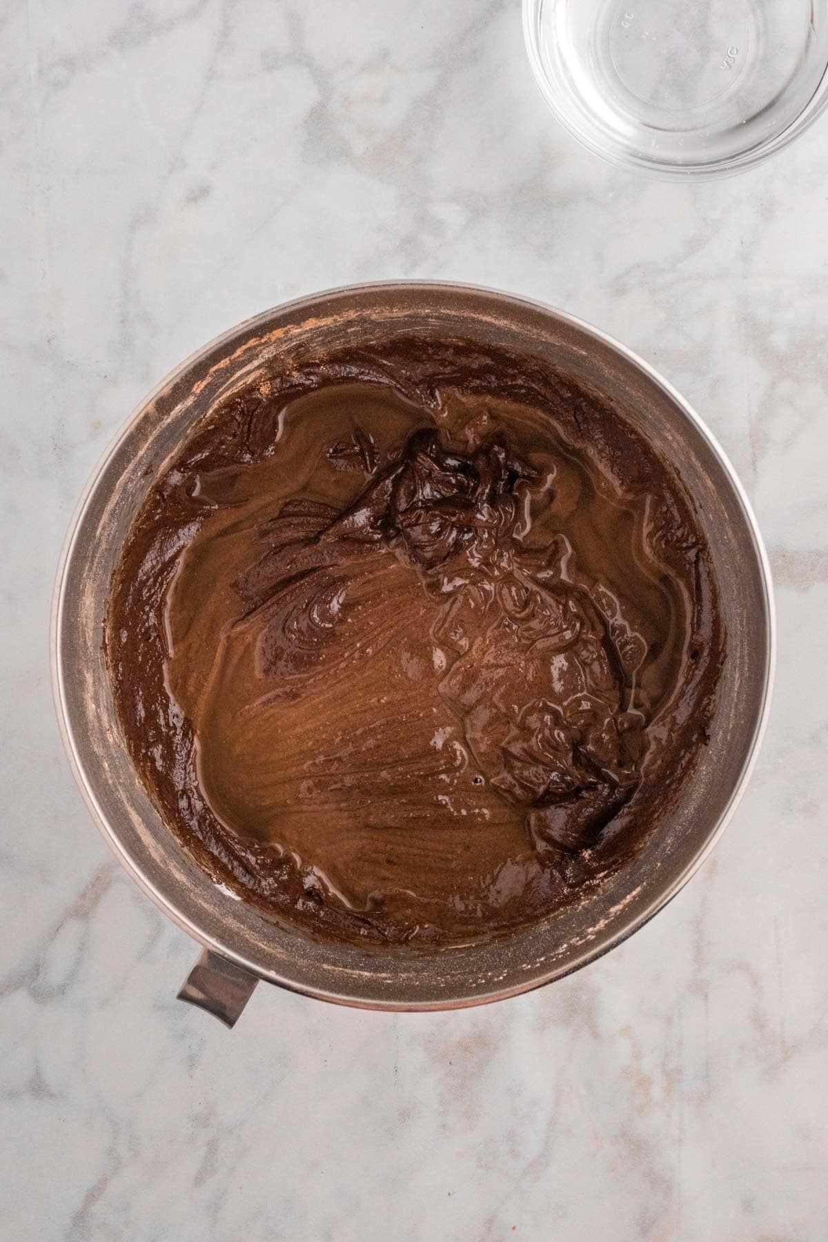 A mixing bowl containing smooth, dark chocolate batter on a marble countertop. A glass bowl is partially visible in the corner.