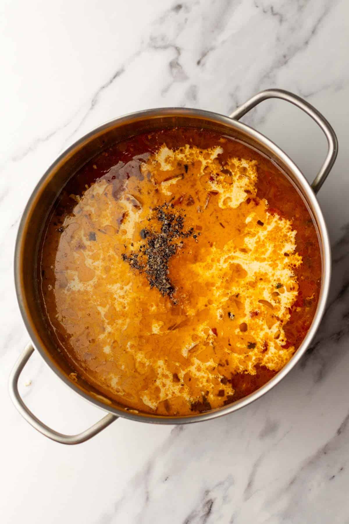 A pot of creamy tomato-based soup with a swirl of cream and sprinkled herbs on a marble countertop.