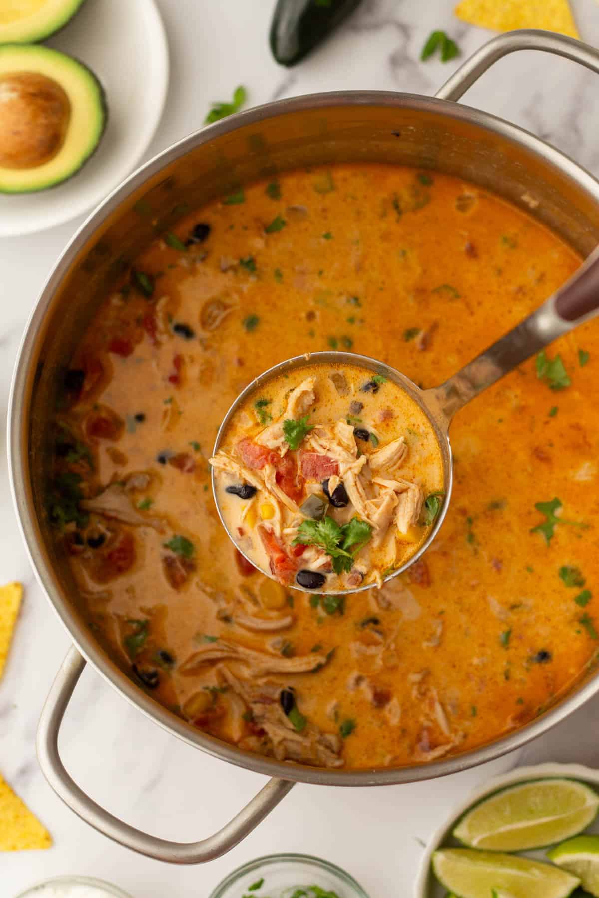 A pot of creamy chicken tortilla soup with tomatoes, black beans, and herbs, with a ladle lifting a portion. Avocado, lime wedges, and tortilla chips surround the pot.