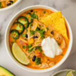 A bowl of creamy chicken tortilla soup topped with sliced jalapeños, tortilla chips, a lime wedge, cheese, and a dollop of sour cream. Nearby are avocado and lime slices.