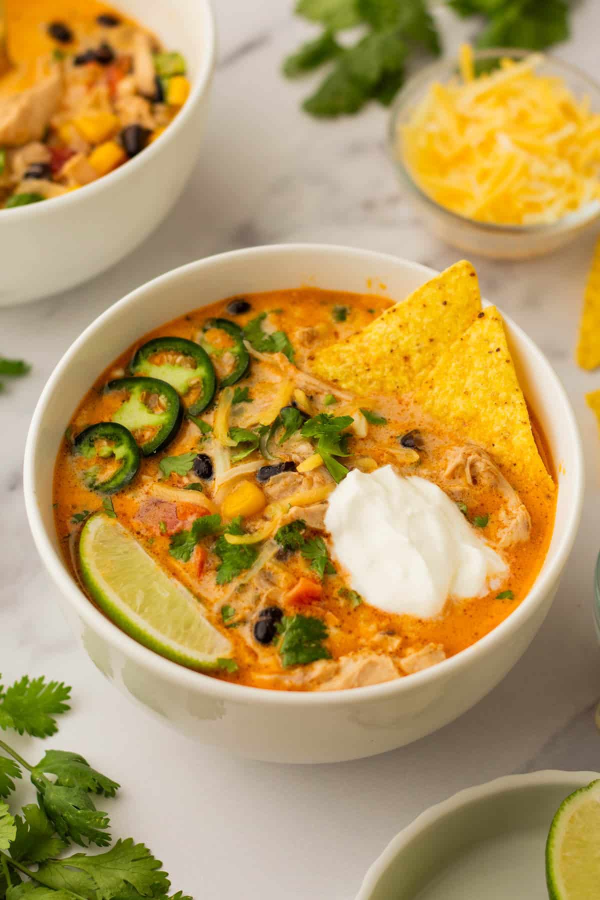 Bowl of creamy chicken tortilla soup with sliced jalape&ntilde;os, tortilla chips, lime wedge, dollop of sour cream, and a sprinkle of cheese and cilantro.