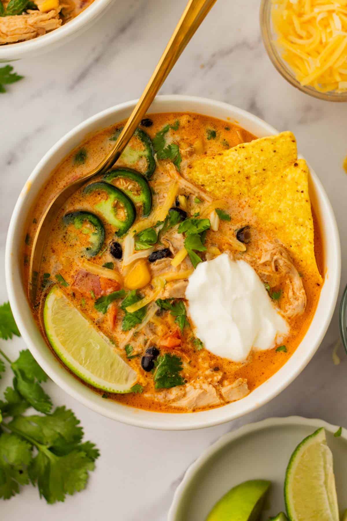 A bowl of creamy chicken tortilla soup topped with sour cream, sliced jalape&ntilde;os, chopped cilantro, and a tortilla chip, with a lime wedge on the side.