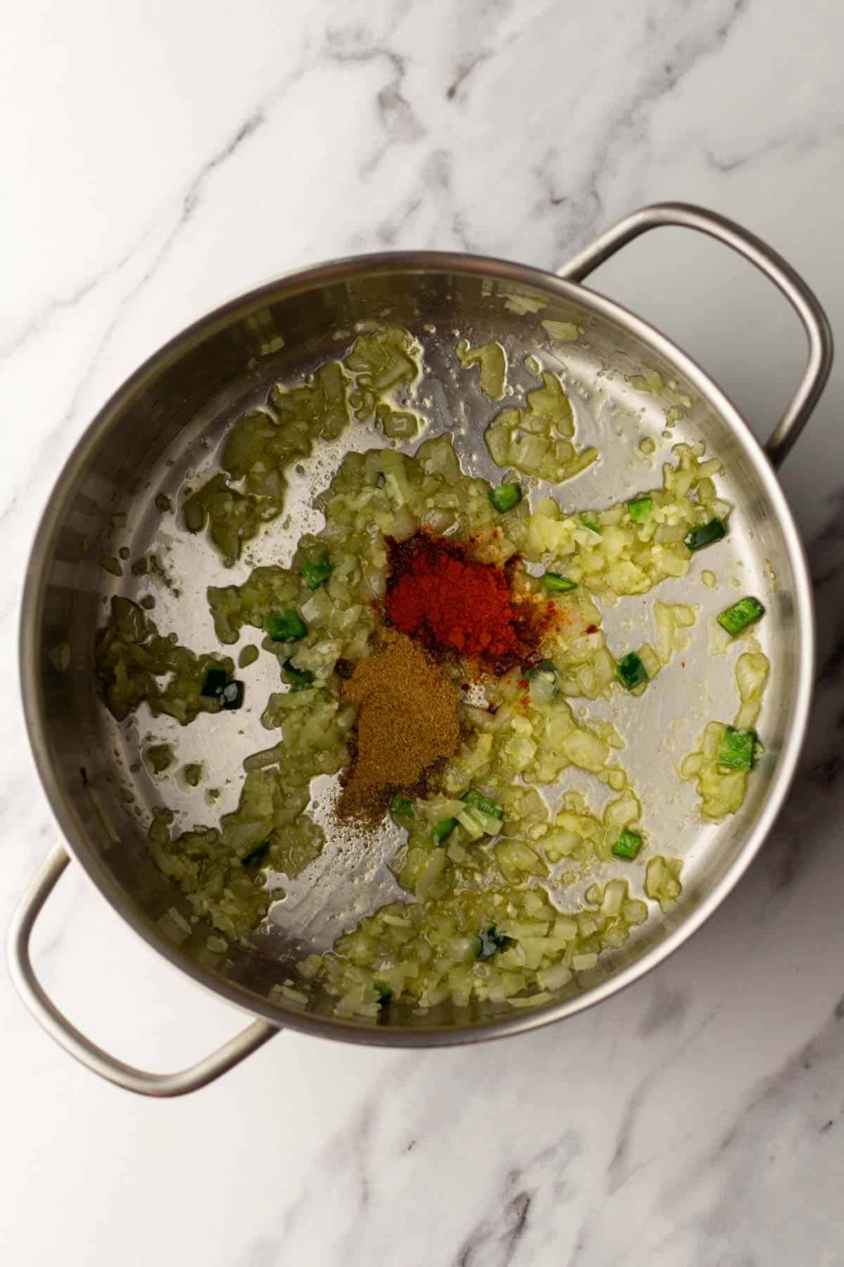 Chopped onions and green peppers saut&eacute;ing in a steel pan with red and brown spices in the center, on a marble countertop.