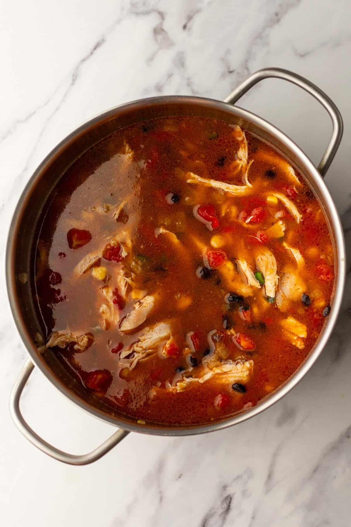 A pot of soup with shredded chicken, black beans, corn, and diced tomatoes on a marble countertop.