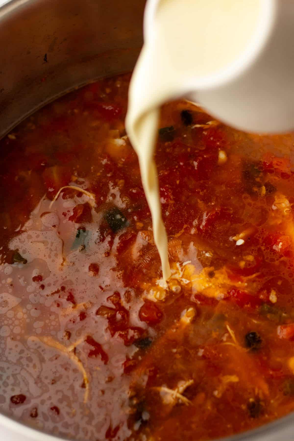 Pouring cream into a pot of tomato-based soup with visible herbs and vegetables.