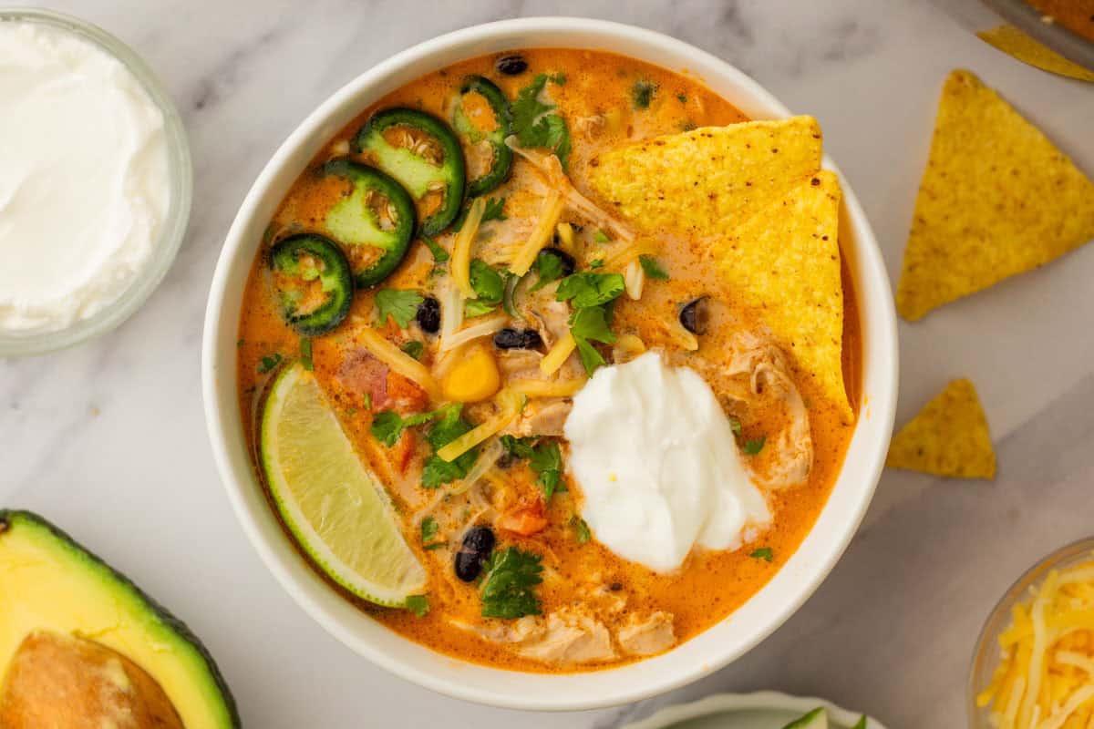 Creamy chicken tortilla soup with black beans, jalape&ntilde;o slices, cheese, sour cream, and a lime wedge, garnished with cilantro. Tortilla chips on the side.