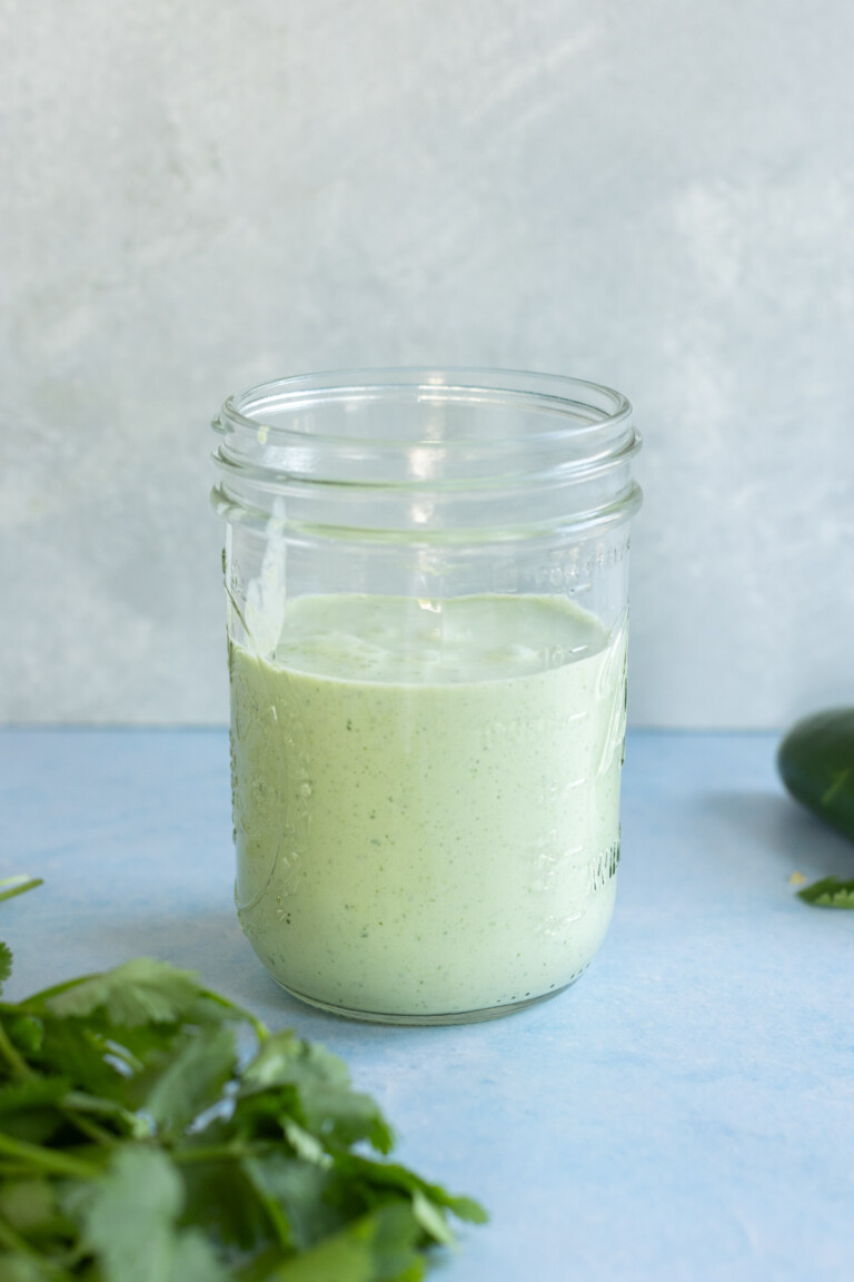 A glass jar filled with jalapeño ranch dressing sits on a blue surface. Fresh green herbs and a darker green vegetable are partially visible nearby.