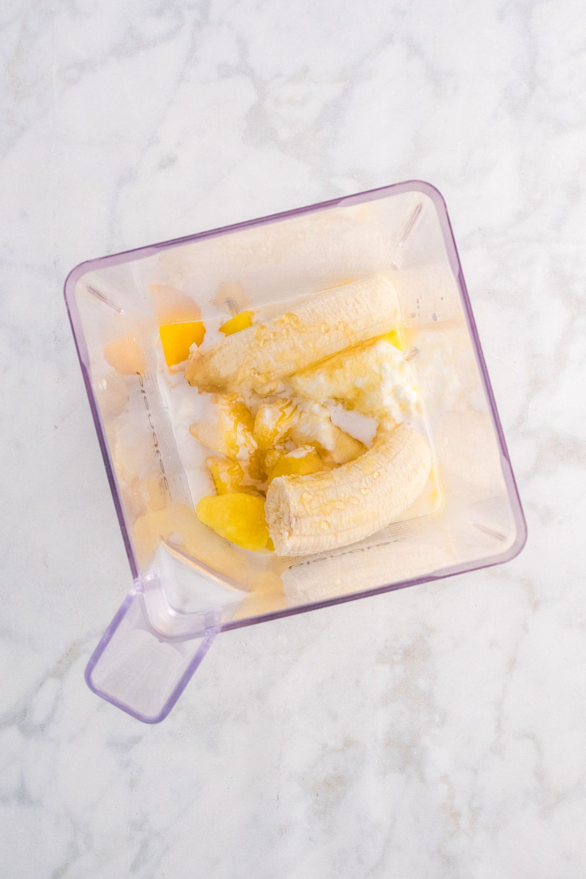 Top view of a blender jar containing peeled banana, mango chunks, yogurt, and honey on a marble countertop.