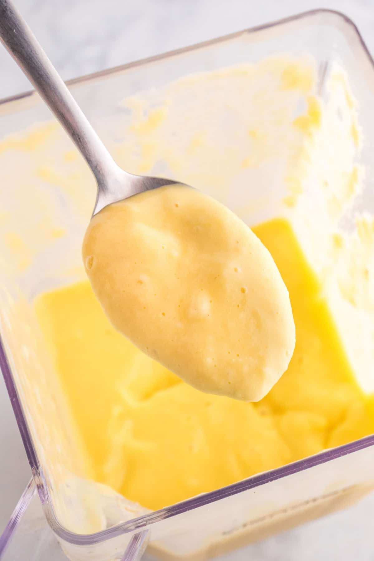 Close-up of a spoon holding a creamy yellow smoothie over a clear container filled with the same mixture.