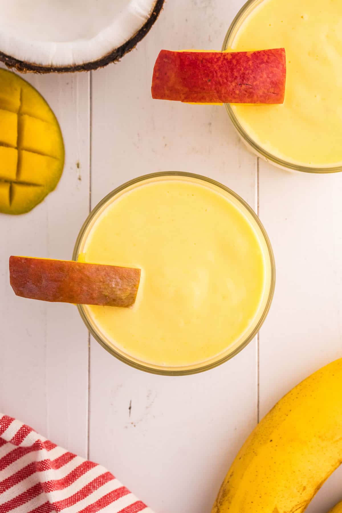 Glasses of a mango pineapple smoothie with apple slices, surrounded by a banana, half a mango, and a coconut shell on a white wooden surface. A red-striped cloth is partially visible.