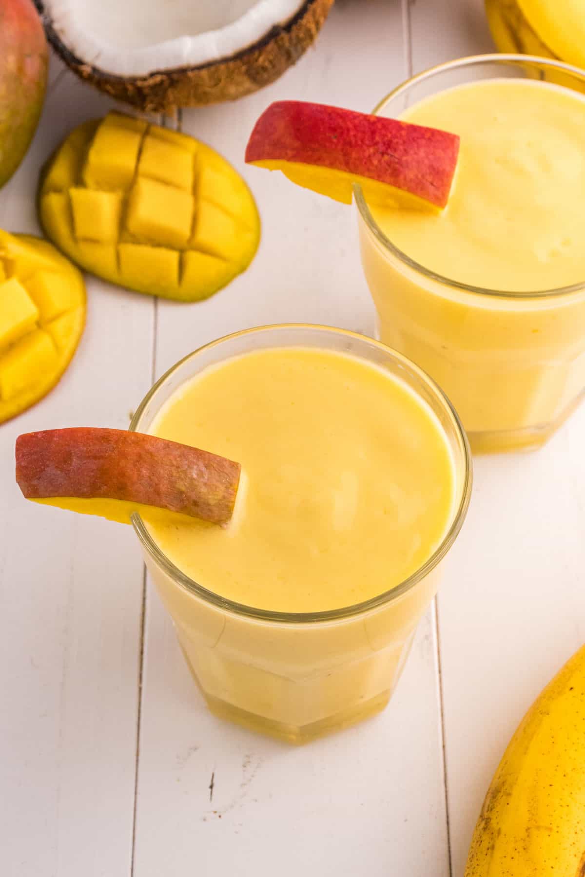 Two mango pineapple smoothies in glasses, each garnished with a peach slice, surrounded by diced and whole mangoes on a white surface.