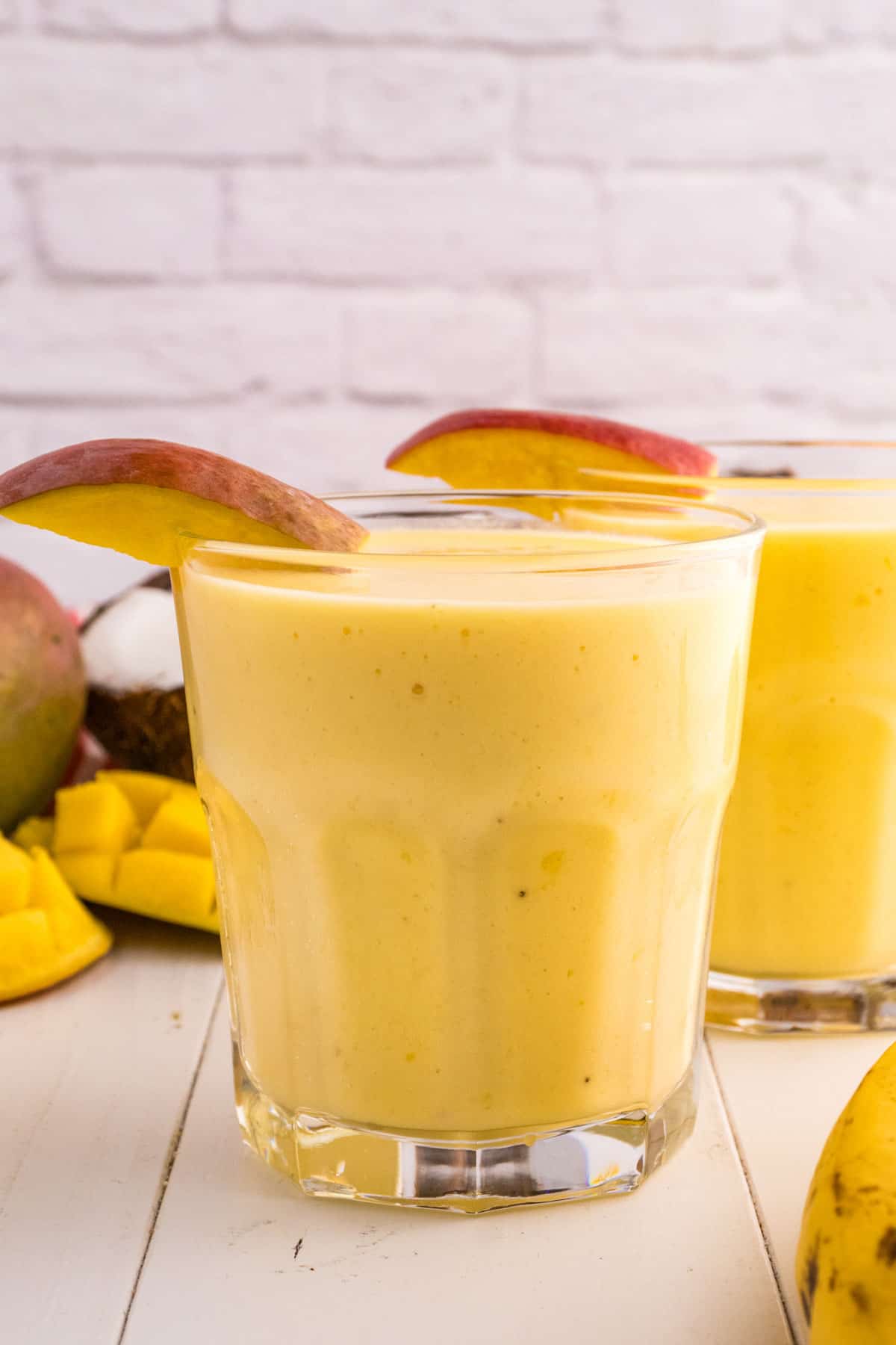 Two glasses of mango pineapple smoothie garnished with mango slices, placed on a white surface with cut mango pieces and whole mangoes in the background.