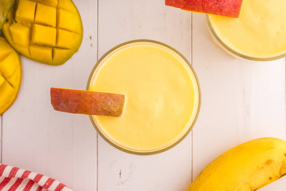 A glass of mango pineapple smoothie topped with a slice of mango on a white wooden surface, surrounded by fresh mango slices and a banana.