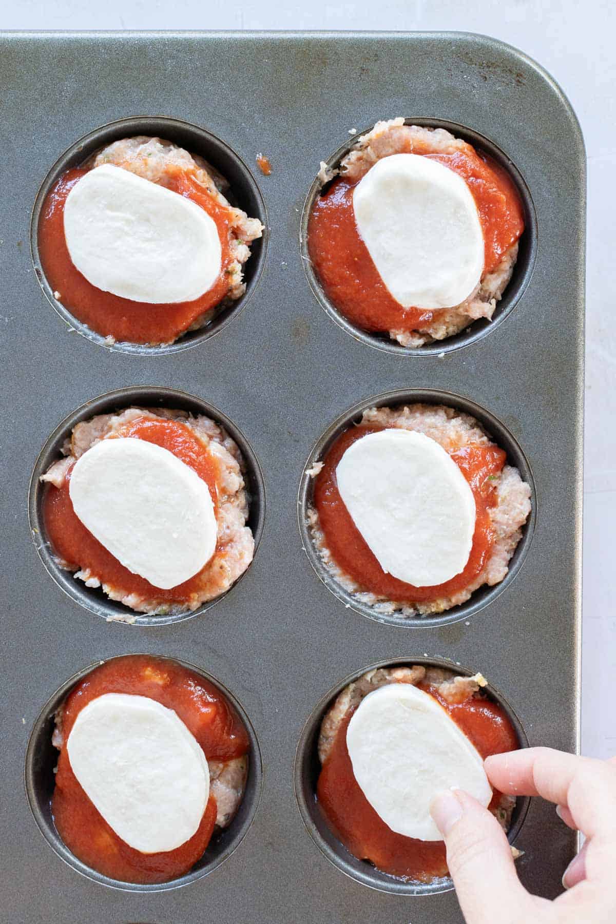 Hand placing mozzarella slices on meatloaf muffins topped with tomato sauce in a muffin tin.