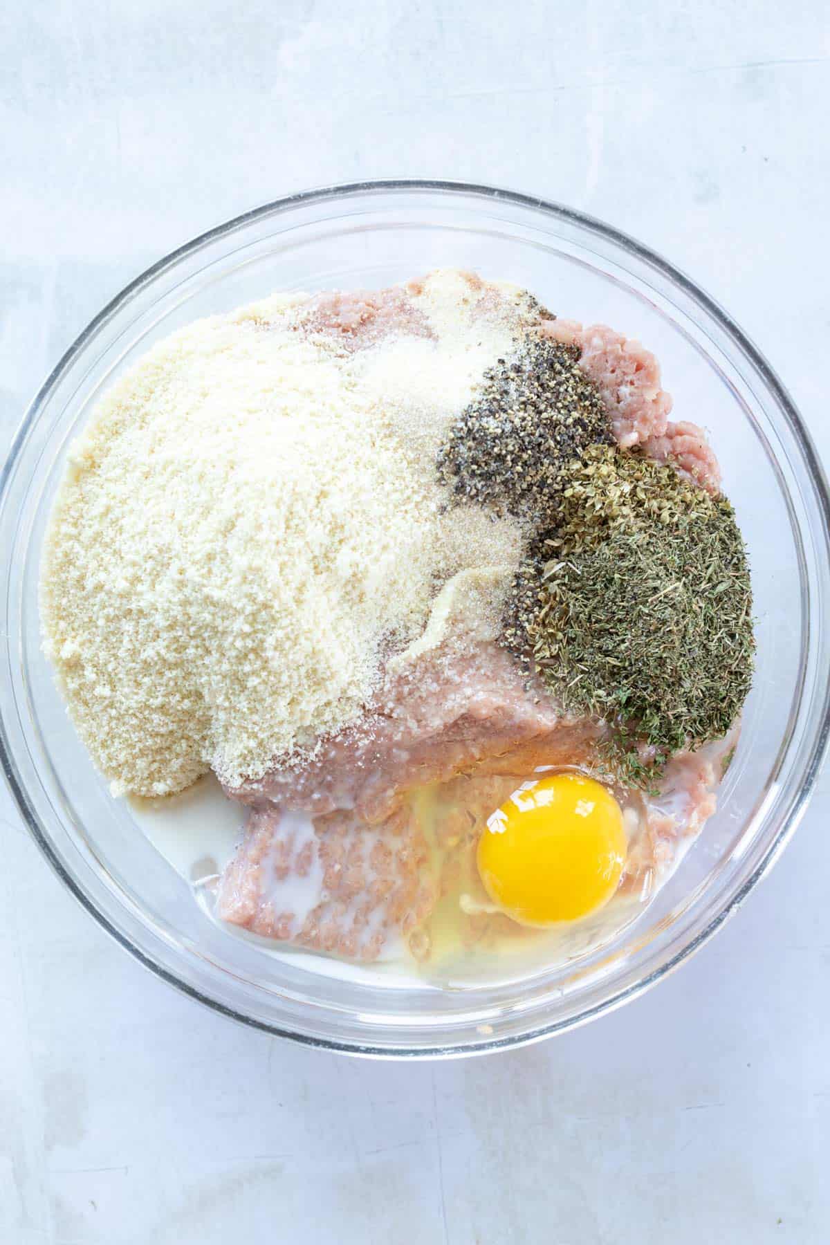 A glass bowl contains ground meat, breadcrumbs, a raw egg, and seasonings, including black pepper and herbs, on a light surface.