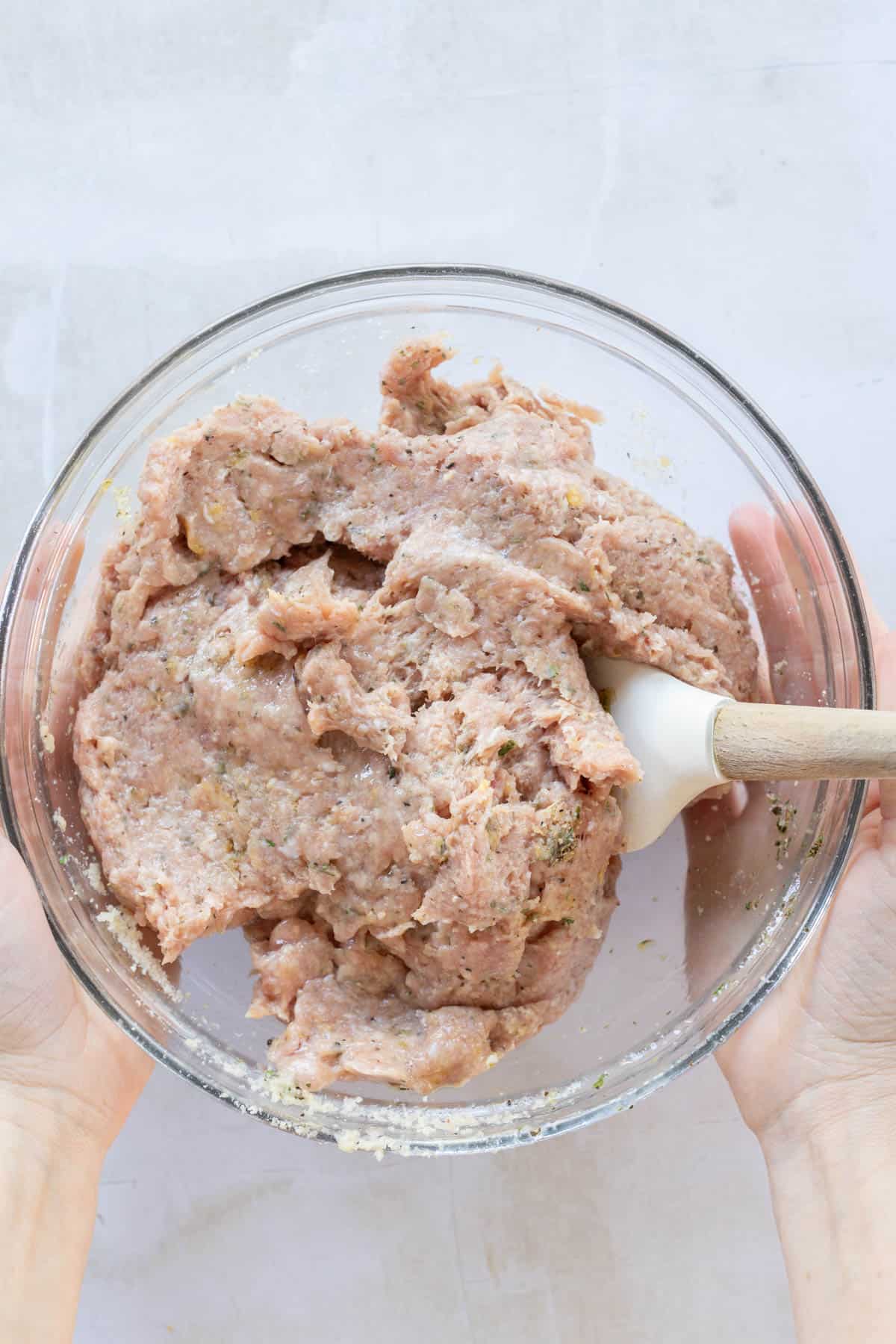 Hands holding a clear glass bowl with raw ground meat mixture being stirred with a white spatula.