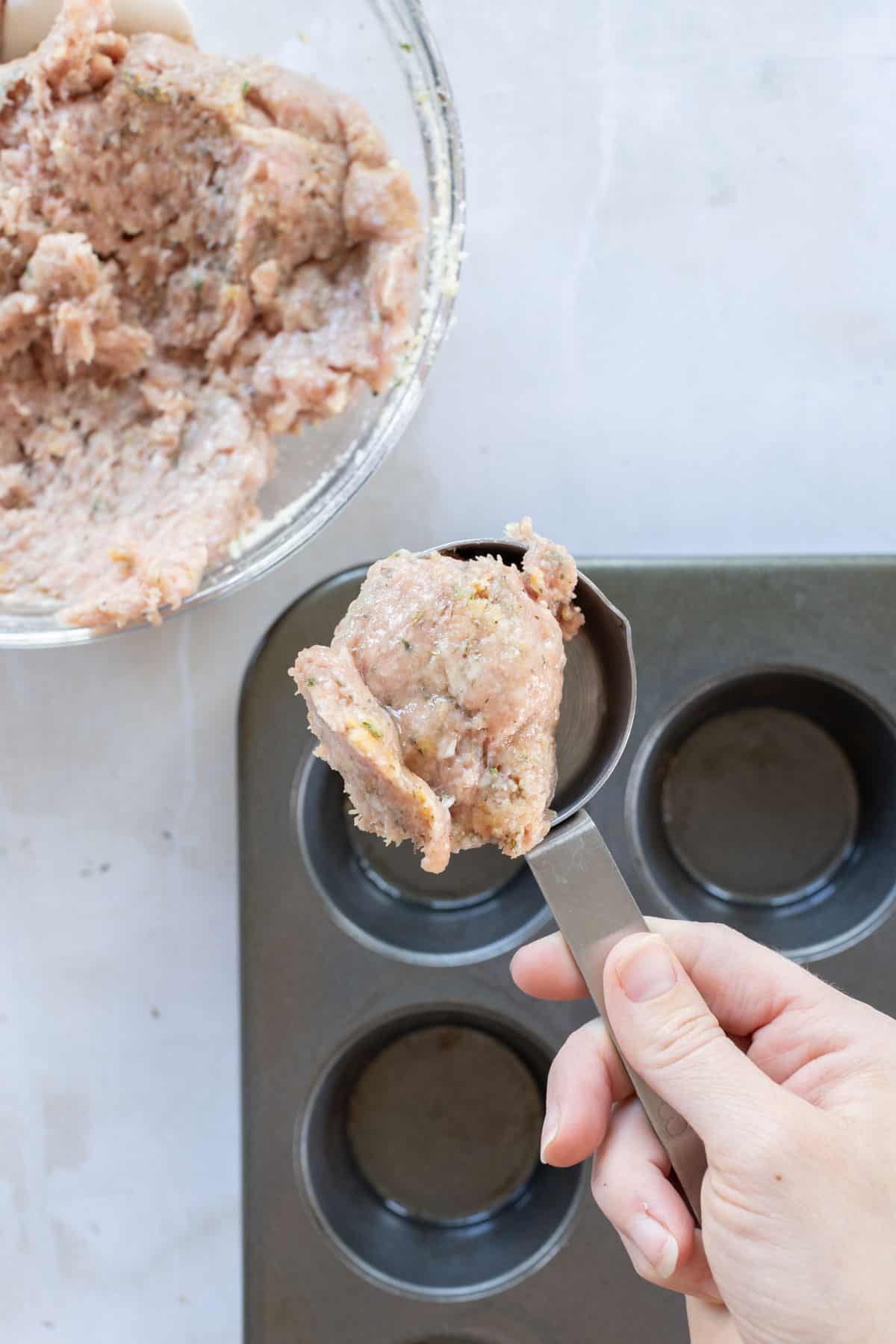 Hand holding a measuring cup with raw minced meat over a muffin tray, with more meat in a bowl nearby.
