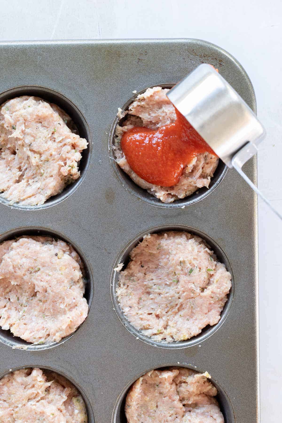 Tomato sauce being poured onto meat mixture in muffin tin cups.