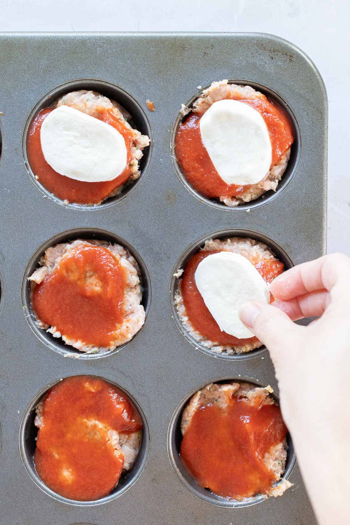 Hand placing mozzarella slices on meatloaf muffins topped with tomato sauce in a muffin tin.