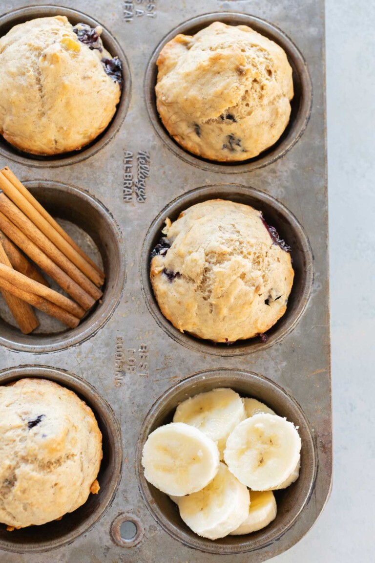 Muffin tin with five baked muffins, slices of banana, and cinnamon sticks. Two muffin slots are empty.