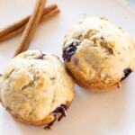 Two blueberry muffins on a white plate with two cinnamon sticks beside them.