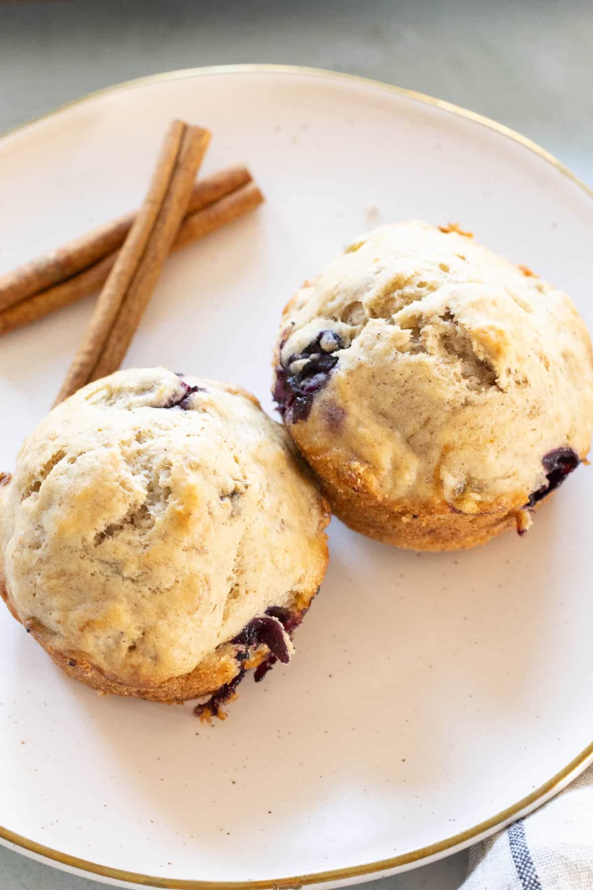 Two blueberry muffins on a white plate with two cinnamon sticks beside them.