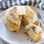 A stack of blueberry pancakes topped with butter on a plate, with a wedge cut out. A fork and a blue-striped cloth are beside the plate.