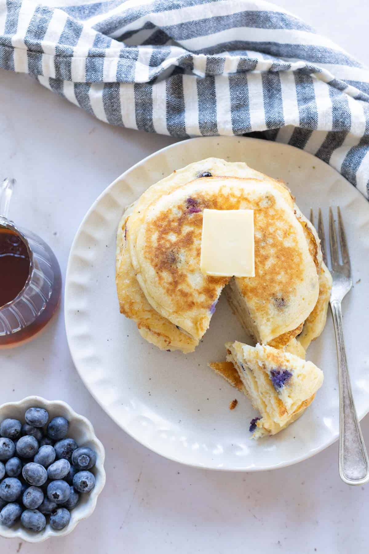 A plate of blueberry pancakes with a pat of butter on top, a fork, blueberries in a bowl, maple syrup in a pitcher, and a striped cloth nearby.