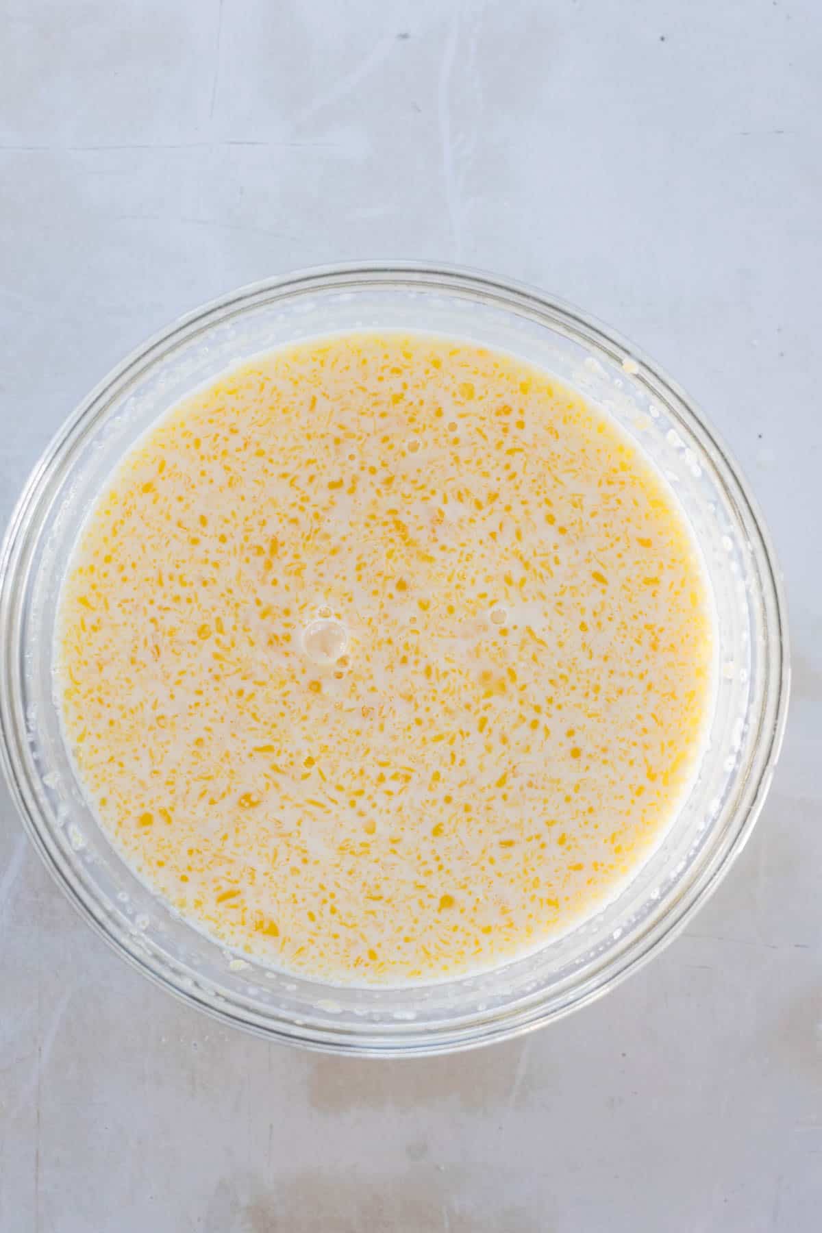 A glass bowl filled with a yellow mixture, resembling beaten eggs, on a light gray surface.