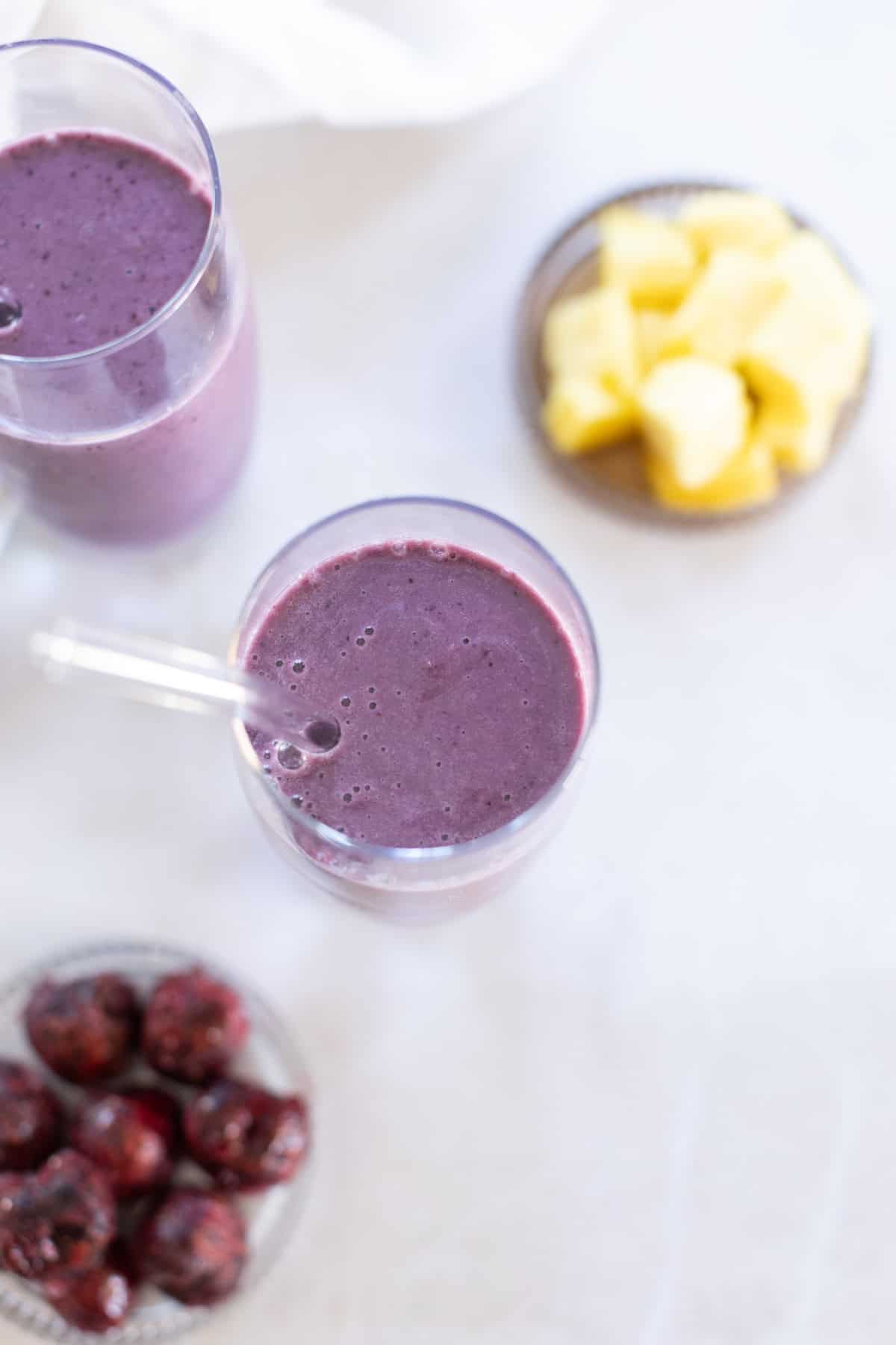 Bird's eye view of two glasses of cherry smoothie with a glass straw, surrounded by bowls of cherries and pineapple chunks on a light surface.
