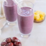 Two glasses of cherry smoothie with clear straws beside a small bowl of dark red fruits and a plate of yellow pineapple chunks on a light surface.