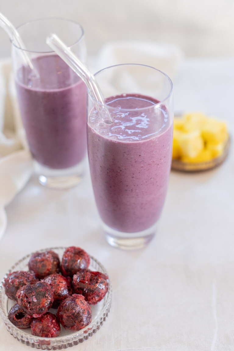 Two glasses of cherry smoothie with clear straws beside a small bowl of dark red fruits and a plate of yellow pineapple chunks on a light surface.