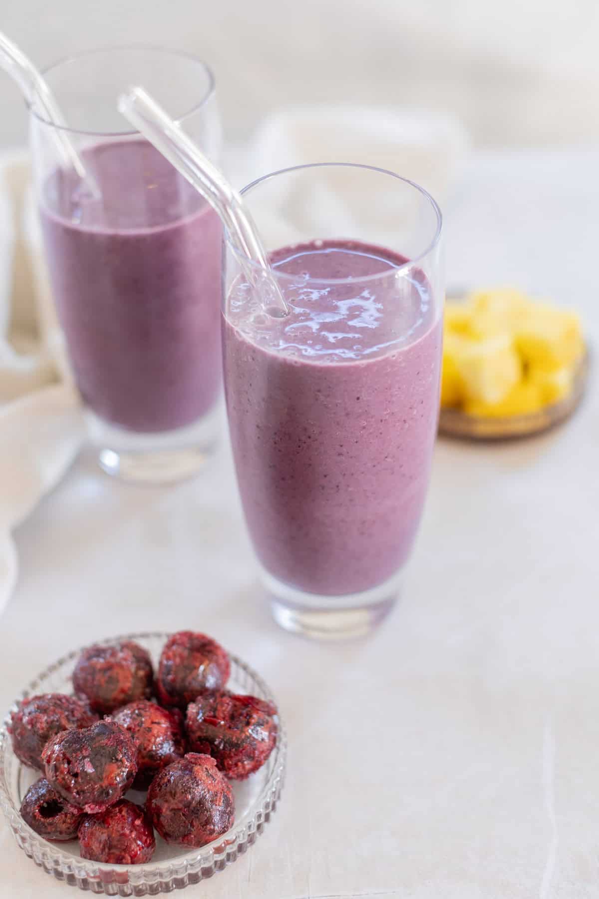 Two glasses of cherry smoothie with clear straws beside a small bowl of dark red fruits and a plate of yellow pineapple chunks on a light surface.