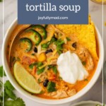 A bowl of creamh chicken tortilla soup topped with jalapeños, lime, cilantro, and sour cream rests on a marble surface, inviting a taste of rich flavors.