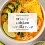A hearty bowl of creamh chicken tortilla soup garnished with lime wedges, jalapeño slices, crunchy tortilla chips, and fresh herbs rests invitingly on a polished marble surface.