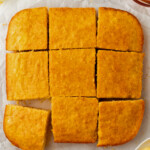 A square pan of cornbread cut into nine pieces, one piece slightly removed. Surrounding items include butter, honey, and a honey dipper.